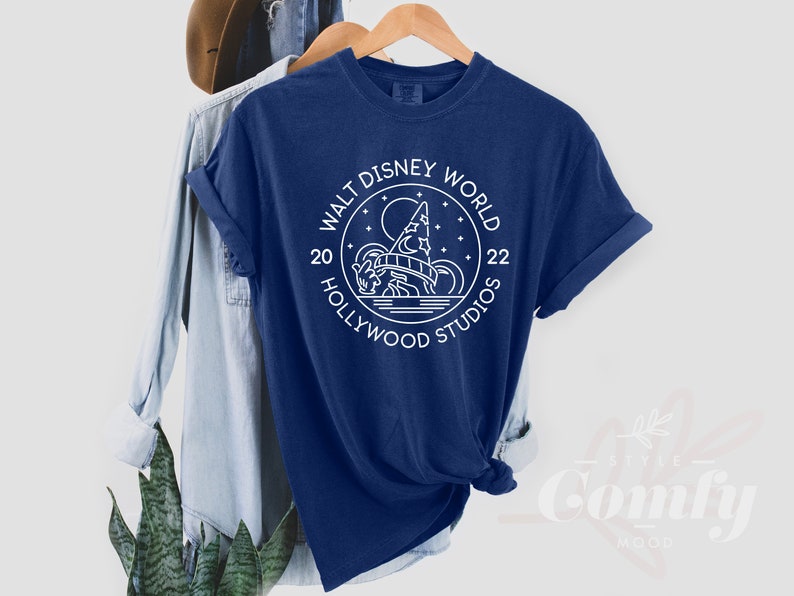 Puede incluir: Una camiseta azul marino con un dise&ntilde;o gr&aacute;fico blanco del logotipo de Walt Disney World Hollywood Studios. El logotipo presenta una imagen estilizada del Hollywood Tower Hotel con una luna y estrellas en el fondo. El texto "Walt Disney World" est&aacute; escrito en un c&iacute;rculo alrededor de la imagen, con "20" y "22" a cada lado del c&iacute;rculo. El texto "Hollywood Studios" est&aacute; escrito debajo del c&iacute;rculo.