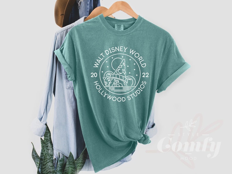 Puede incluir: Una camiseta azul turquesa con un dise&ntilde;o gr&aacute;fico blanco en forma de c&iacute;rculo con el texto "Walt Disney World 20 Hollywood Studios 22" y una ilustraci&oacute;n estilizada del Hollywood Tower Hotel.