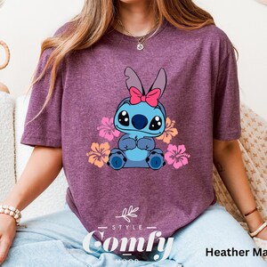 May include: A heather maroon t-shirt featuring a cartoon Stitch character with pink flowers and a pink bow. The shirt has the text "Comfy" in a white font with a black outline.