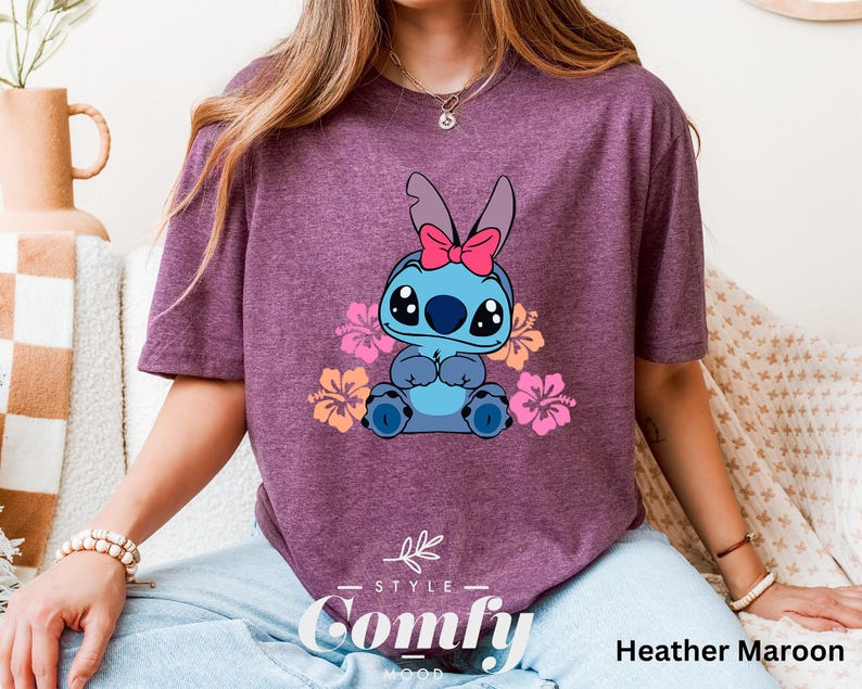May include: A heather maroon t-shirt featuring a cartoon Stitch character with pink flowers and a pink bow. The shirt has the text "Comfy" in a white font with a black outline.