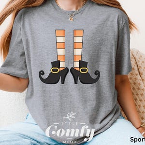 May include: Sport grey t-shirt featuring a whimsical graphic of witch legs with orange and white striped stockings and black shoes with gold buckles. The text "Style Comfy Mood" is printed at the bottom.
