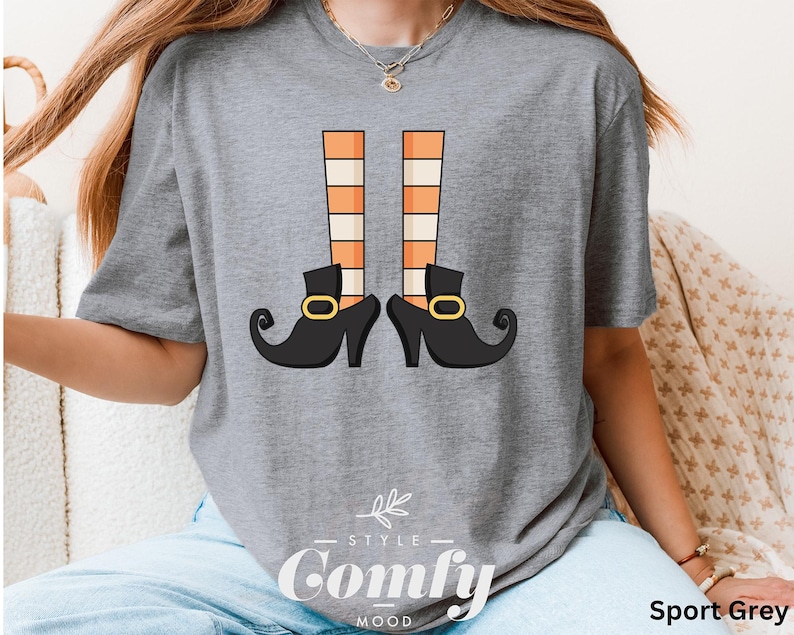 May include: Sport grey t-shirt featuring a whimsical graphic of witch legs with orange and white striped stockings and black shoes with gold buckles. The text "Style Comfy Mood" is printed at the bottom.