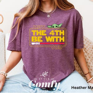May include: A heather maroon t-shirt with a yellow graphic that says "May the 4th be with you" and a cartoon image of a green alien. The graphic is in a yellow rectangle with a red line across the top. The shirt is from the brand "Comfy".