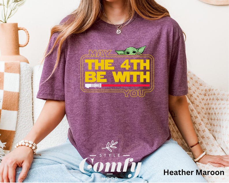 May include: A heather maroon t-shirt with a yellow graphic that says "May the 4th be with you" and a cartoon image of a green alien. The graphic is in a yellow rectangle with a red line across the top. The shirt is from the brand "Comfy".