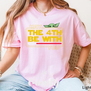 May include: A light pink t-shirt with a graphic of a green alien peeking out of a box with the text "May the 4th be with you".
