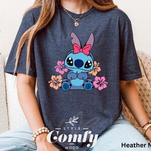 May include: A heather navy blue t-shirt featuring a cartoon Stitch character with pink flowers. The shirt has the text "Comfy" printed on the bottom.