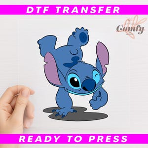 May include: A DTF transfer featuring a cartoon character in shades of blue with large eyes and ears. The character is winking and appears to be walking. The text "DTF TRANSFER" and "READY TO PRESS" are displayed in pink.