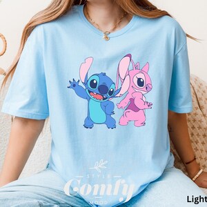 May include: Light blue t-shirt featuring a cartoon graphic of Stitch and Angel. The blue Stitch is waving, and the pink Angel is walking. The text "STYLE" and "Comfy Mood" is printed at the bottom of the shirt. The words "Light Blue" are printed on the bottom right.