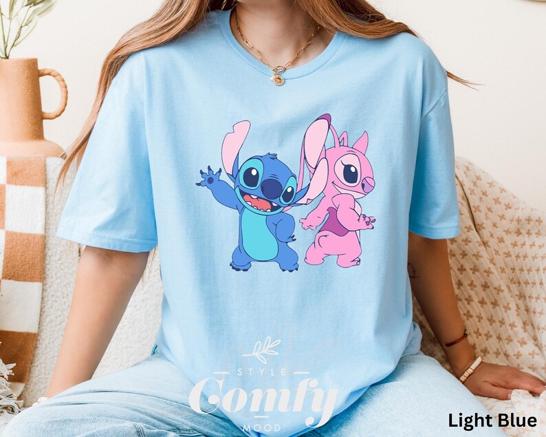 May include: Light blue t-shirt featuring a cartoon graphic of Stitch and Angel. The blue Stitch is waving, and the pink Angel is walking. The text "STYLE" and "Comfy Mood" is printed at the bottom of the shirt. The words "Light Blue" are printed on the bottom right.