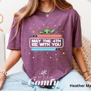 K&ouml;nnte beinhalten: Ein T-Shirt in Heide-Maroon mit einem Motiv eines gr&uuml;nen Au&szlig;erirdischen, der hinter einem Lichtschwert hervorlugt. Der Text "May the 4th Be With You" ist auf dem T-Shirt aufgedruckt.