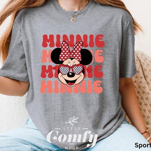 Puede incluir: Una camiseta gris con un gr&aacute;fico de Minnie Mouse rojo y blanco y el texto "Minnie" repetido tres veces en una fuente rosa y blanca. La camiseta tiene cuello redondo y mangas cortas. El texto "Comfy" est&aacute; impreso en la parte inferior de la camiseta en una fuente negra.