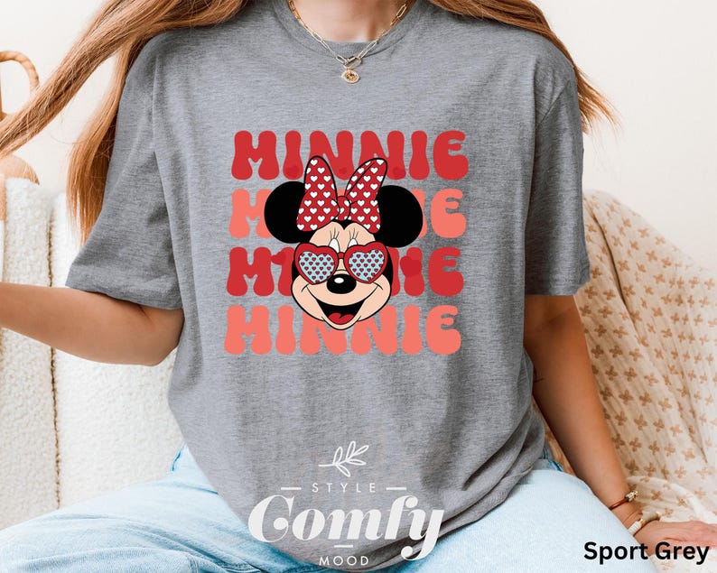 Puede incluir: Una camiseta gris con un gr&aacute;fico de Minnie Mouse rojo y blanco y el texto "Minnie" repetido tres veces en una fuente rosa y blanca. La camiseta tiene cuello redondo y mangas cortas. El texto "Comfy" est&aacute; impreso en la parte inferior de la camiseta en una fuente negra.