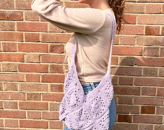 May include: A light purple crochet shoulder bag with a geometric pattern. The bag is worn over the shoulder. The background is a red brick wall.