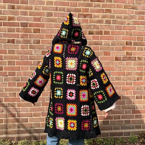 May include: A black hooded crochet coat featuring colorful square patterns. The coat has long sleeves and a hood, with a variety of bright colors in the squares, including pink, yellow, green, and blue. The coat is worn over a white shirt and blue jeans.