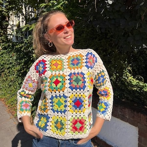 Könnte beinhalten: Eine Frau trägt ein weißes Häkeltop mit bunten Quadraten. Das Top hat einen quadratischen Ausschnitt und Dreiviertelärmel. Sie trägt eine orangefarbene Sonnenbrille und hat ihre Haare zu einem Pferdeschwanz gebunden.