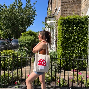 Pu&ograve; includere: Una borsa tote bianca con un designo a forma di fungo rosso e bianco. La borsa &egrave; portata da una persona con una canotta marrone e pantaloncini di jeans.