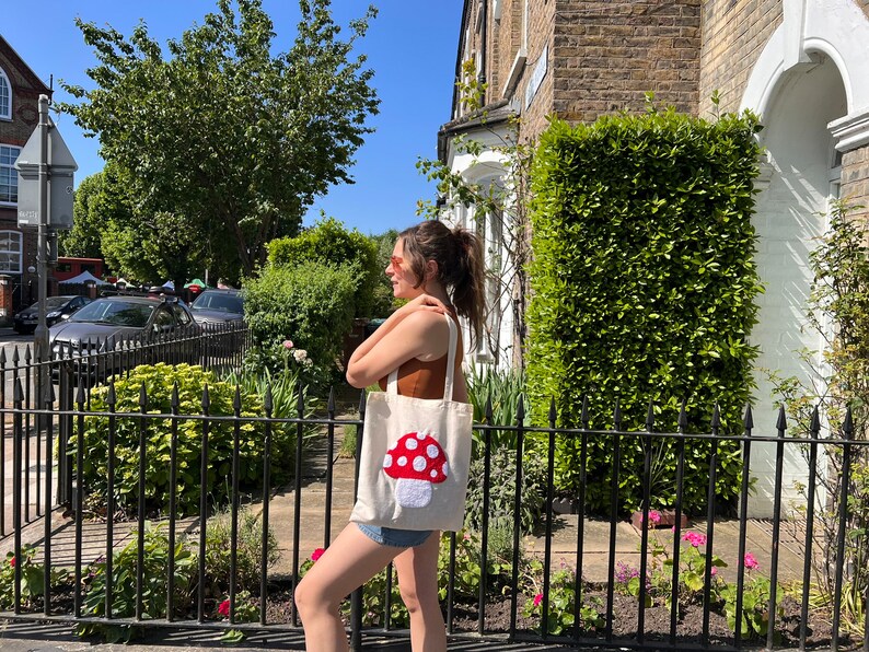 Pu&ograve; includere: Una borsa tote bianca con un designo a forma di fungo rosso e bianco. La borsa &egrave; portata da una persona con una canotta marrone e pantaloncini di jeans.