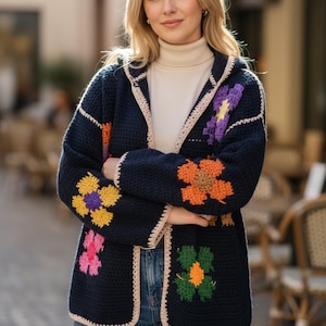 以下が含まれることがあります： ネイビーブルーのかぎ針編みのカーディガン。フード付きで、黄色、オレンジ、紫、ピンクのカラフルな花のアップリケが施されています。カーディガンはベージュのトリムで、クリーム色のタートルネックとブルージーンズと合わせています。
