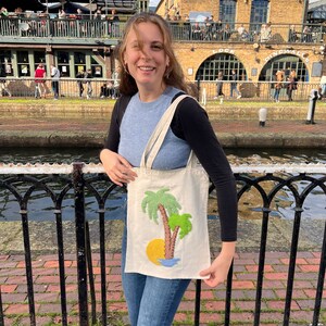 May include: A person is holding a white tote bag with an embroidered design of two palm trees and a yellow sun. The bag is in front of a brick building with the text "The  T.E. Dingwall Building" on the side.