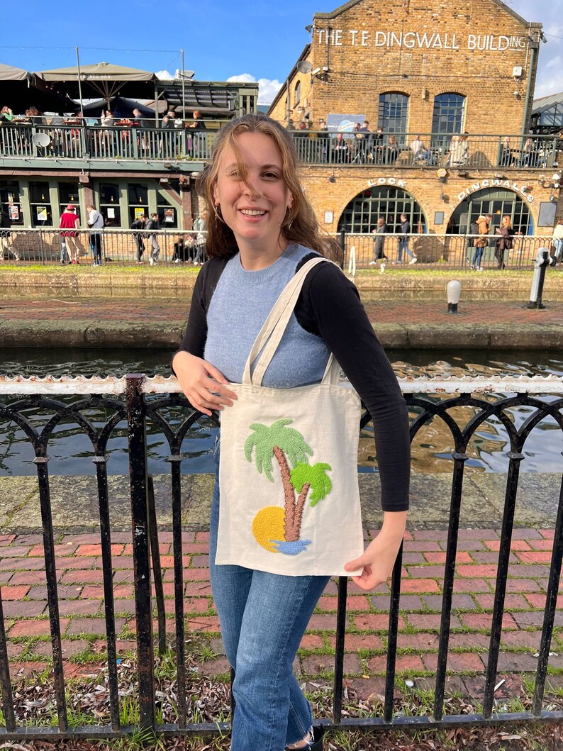 May include: A person is holding a white tote bag with an embroidered design of two palm trees and a yellow sun. The bag is in front of a brick building with the text "The  T.E. Dingwall Building" on the side.