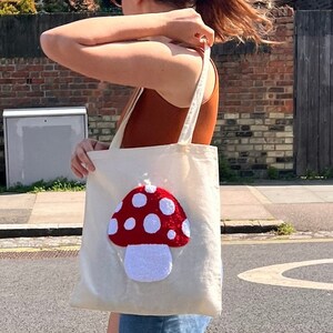 Pu&ograve; includere: Una borsa tote bianca con un designo a forma di fungo rosso e bianco. Il fungo &egrave; fatto di tessuto soffice e ha dei puntini bianchi sul cappello rosso.