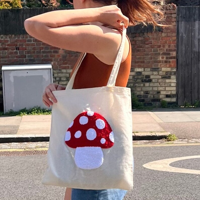Pu&ograve; includere: Una borsa tote bianca con un designo a forma di fungo rosso e bianco. Il fungo &egrave; fatto di tessuto soffice e ha dei puntini bianchi sul cappello rosso.