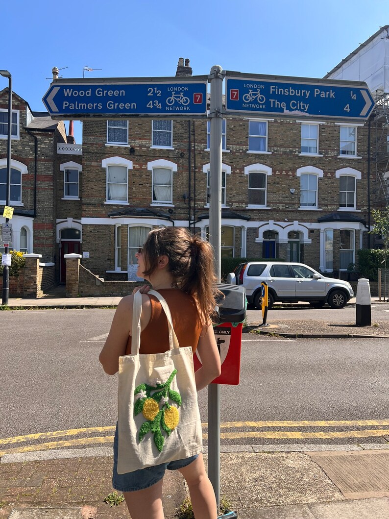 Pu&ograve; includere: Una donna &egrave; in piedi su un marciapiede di fronte a una fila di edifici in mattoni. Porta una borsa tote bianca con un design di limone ricamato. La borsa ha una tracolla marrone. Ci sono cartelli stradali sullo sfondo che indicano "Wood Green, Palmers Green, Finsbury Park, The City".