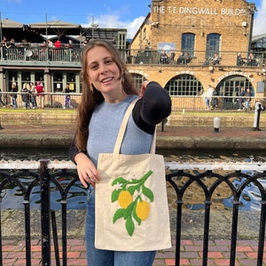 May include: A woman is holding a white tote bag with an embroidered lemon branch design. The bag has green leaves and yellow lemons. The woman is standing in front of a brick building with the text "The Teddingwall Building" on it.