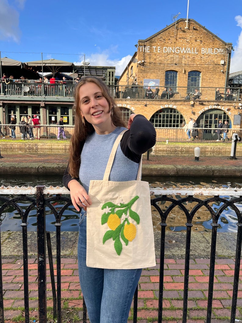 May include: A woman is holding a white tote bag with an embroidered lemon branch design. The bag has green leaves and yellow lemons. The woman is standing in front of a brick building with the text "The Teddingwall Building" on it.