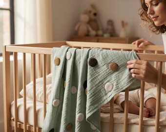 May include: A light green baby blanket with large, textured, circular accents in various colors, draped over a wooden crib. The crib has a light-colored mattress and a person is seen near the crib.
