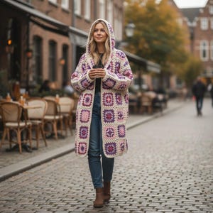 May include: A long, hooded crochet cardigan in a granny square pattern. The open-front sweater features squares of cream, pink, and purple. The woman is wearing blue jeans and brown boots. The background shows a cobblestone street and buildings.