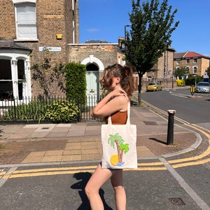 Pu&ograve; includere: Una persona cammina per strada portando una borsa di tela bianca con un disegno di palma verde. La borsa ha un sole giallo e acqua blu ricamata.