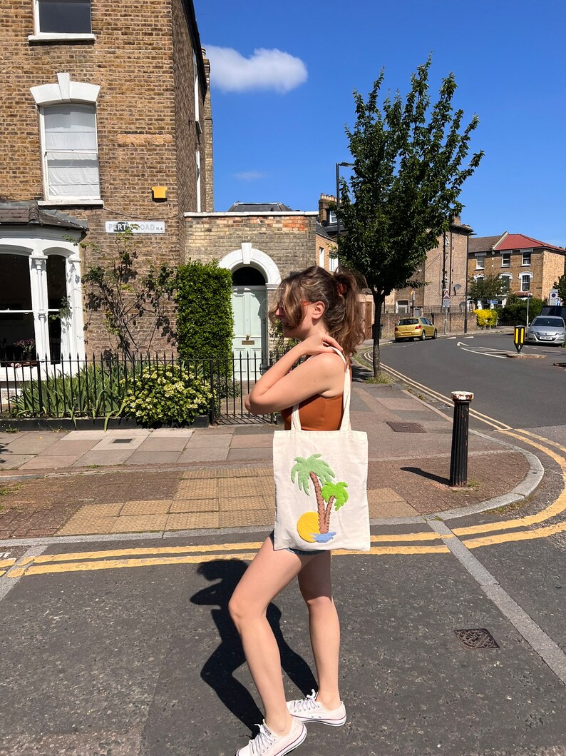 Pu&ograve; includere: Una persona cammina per strada portando una borsa di tela bianca con un disegno di palma verde. La borsa ha un sole giallo e acqua blu ricamata.