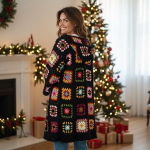 Könnte beinhalten: Ein schwarzer Kapuzen-Häkelcardigan mit bunten quadratischen Mustern. Der langärmlige Pullover hat ein Patchwork-Design mit verschiedenen Farben, darunter Rosa, Gelb, Blau und Grün. Der Cardigan wird vor einem Weihnachtsbaum getragen.
