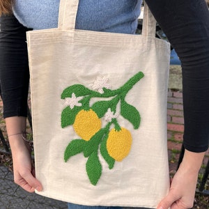 May include: A white canvas tote bag with a green and yellow embroidered lemon tree design. The tote bag has two handles.
