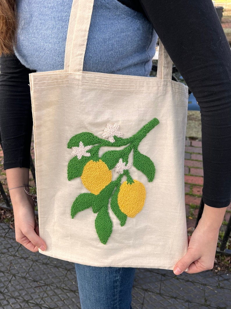 May include: A white canvas tote bag with a green and yellow embroidered lemon tree design. The tote bag has two handles.
