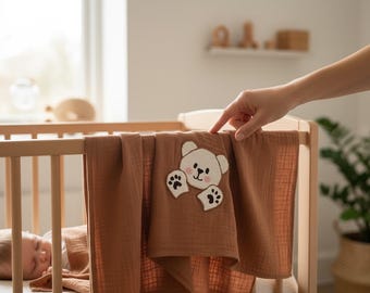 May include: A brown baby blanket with a white teddy bear applique on a wooden crib. The bear has black paw prints and a pink nose. A person's hand is touching the blanket.