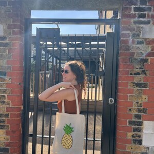 Pu&ograve; includere: Una persona porta una borsa tote bianca con un disegno di ananas giallo. La borsa &egrave; appesa alla sua spalla. La persona &egrave; in piedi davanti a un muro di mattoni con un cancello di metallo su cui &egrave; scritto "GIRLS".