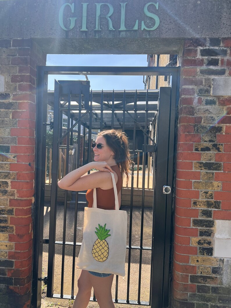 Pu&ograve; includere: Una persona porta una borsa tote bianca con un disegno di ananas giallo. La borsa &egrave; appesa alla sua spalla. La persona &egrave; in piedi davanti a un muro di mattoni con un cancello di metallo su cui &egrave; scritto "GIRLS".