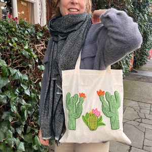 May include: A white canvas tote bag with a green, orange, and pink embroidered cactus design. The bag is being held by a person wearing a gray sweater and a black and white scarf.