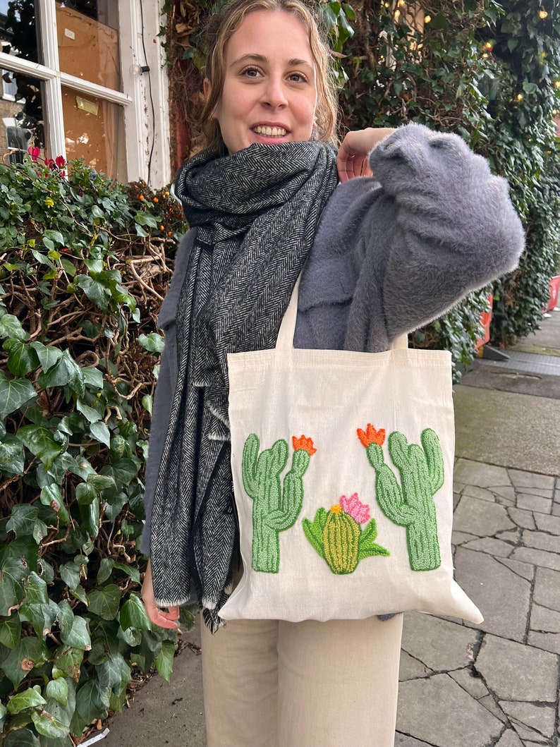 May include: A white canvas tote bag with a green, orange, and pink embroidered cactus design. The bag is being held by a person wearing a gray sweater and a black and white scarf.