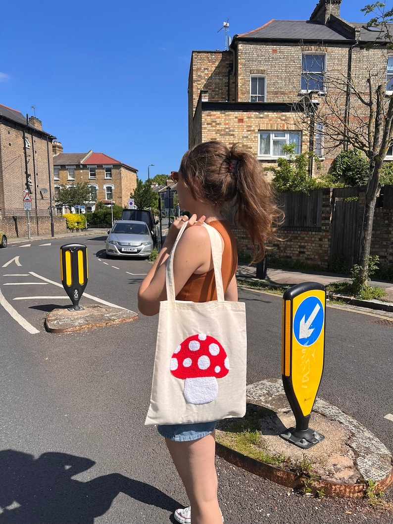 Pu&ograve; includere: Una borsa tote bianca con un designo a forma di fungo rosso e bianco. Il fungo &egrave; ricamato con filo bianco e ha punti rossi in feltro.