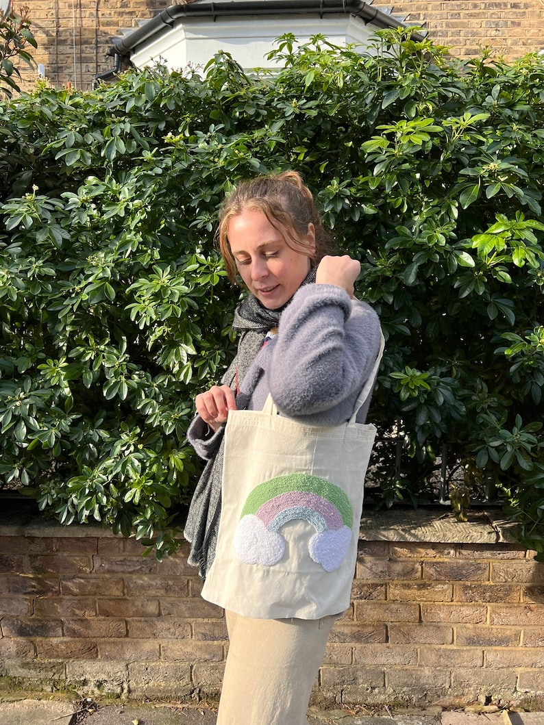 May include: A white canvas tote bag with a fluffy rainbow and cloud design. The rainbow is green, light blue, and pink.