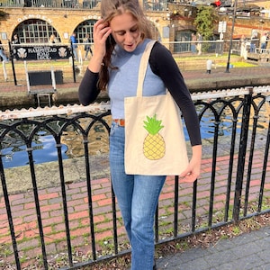 May include: A woman is holding a beige tote bag with a pineapple design. The pineapple is yellow with green leaves. The bag has a long strap and is worn over the shoulder. The woman is wearing a blue tank top and jeans. She is standing in front of a black metal fence.