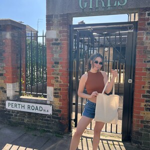 Pu&ograve; includere: Una persona indossa un top marrone, pantaloncini di jeans e scarpe da ginnastica bianche e si trova davanti a un edificio in mattoni con un cancello sopra il quale &egrave; scritto "GIRLS". La persona tiene in mano una borsa tote beige. Un cartello con la scritta "PERTH ROAD N.4" &egrave; appeso al muro.