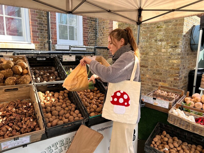 May include: A woman at a farmers market is holding a white tote bag with a red and white mushroom design. The bag is filled with produce. The tote bag has the text "Infuse Yourself" printed on it.