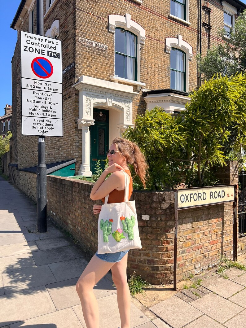 Pu&ograve; includere: Una donna cammina lungo una strada di fronte a un edificio in mattoni con un cartello che recita "Finsbury Park C Controlled ZONE FPC". Porta una borsa di tela bianca con un cactus verde ricamato. Il cartello contiene anche informazioni sulle restrizioni di parcheggio.