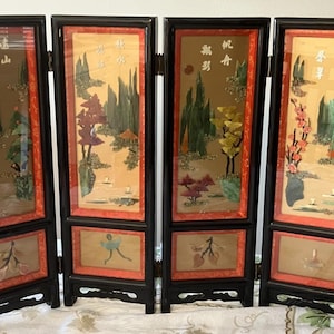 May include: A four-panel folding screen with a black frame and red borders. Each panel features a landscape scene with raised, colorful stone-like elements and Chinese characters. The lower sections contain smaller, separate rectangular insets.