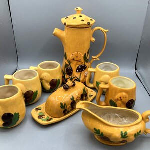 Op de afbeelding: Een geel keramieken theeservies bestaande uit een hoge theepot met een paddenstoelvormig deksel, vier mokken, een botervloot met deksel en een sauskom. Elk item is versierd met geschilderde bruine paddenstoelen en groene bladeren.