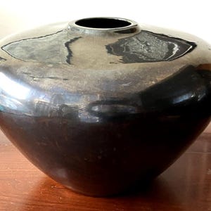 May include: A wide, low-profile vase with a smooth, highly reflective black finish. The vessel features a rounded, bulbous body that tapers toward a small, circular opening at the top, creating a sleek, continuous silhouette with deep dark tones.